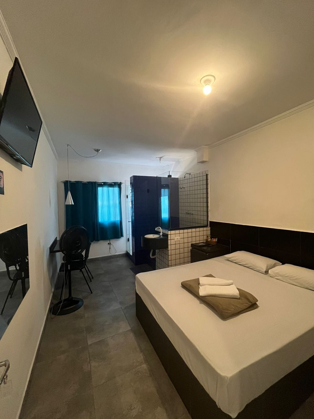 Quarto de hotel com cama, escrivaninha, cadeira, televisão e banheiro.