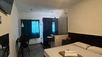 Quarto de hotel com cama, escrivaninha, cadeira, televisão e banheiro.