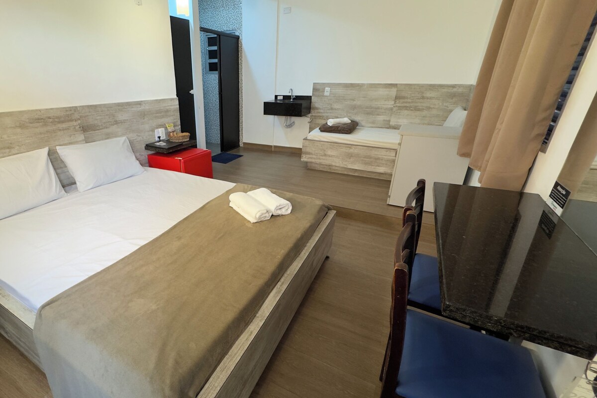 A hotel room with a bed, a desk, a chair, a small table, a sink, and a mirror.