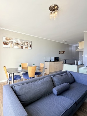 Una sala de estar moderna con un sofá gris, una mesa de comedor con sillas, una zona de cocina y una lámpara en el techo.