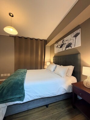 Una cama bien hecha con un cabecero gris, una lámpara de noche y una obra de arte colgada en la pared.