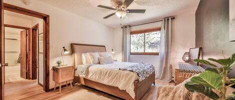 Un dormitorio con cama, mesitas de noche, ventilador de techo, ventana con cortinas y vistas a árboles.