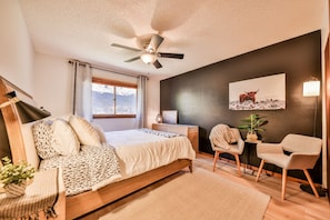 A bedroom with a bed, a desk, two chairs, a ceiling fan, a window, and a wall-mounted lamp.