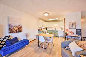 A modern living room with a sofa, a dining table with chairs, a kitchen area with cabinets, and a mounted artwork.
