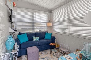 Ein gemütliches Wohnzimmer mit einem dunkelblauen Sofa, einem Glastisch, einem gemusterten Teppich und einem großen Fenster mit Jalousien.