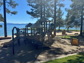 Ein Spielplatz mit Rutsche und Klettergerüsten, umgeben von Bäumen und Blick auf einen See.
