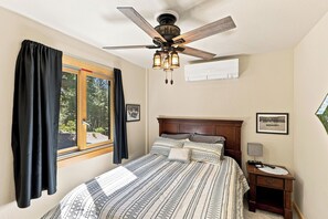 Ein Schlafzimmer mit einem Bett, einem Nachttisch, einer Wand-Klimaanlage, einem Deckenventilator und einem fensterbelaubten Fenster.