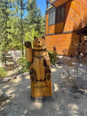 Eine Holzskulptur eines Bären, deren Sockel mit 'WELCOME' beschriftet ist, steht in einer Schotterfläche mit Bäumen und einem Holzgebäude im Hintergrund.