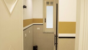 Ein schmaler Gang mit einer Tür links, einem Fenster mit satiniertem Glas und einem schwarzen Mülleimer auf dem Boden.