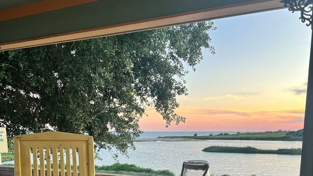 Une terrasse en bois avec un banc et une table, donnant sur une étendue d’eau et offrant une vue sur le coucher de soleil.
