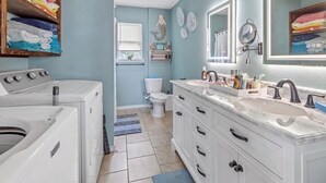 Une salle de bain équipée d’un lave-linge, de toilettes, d’un meuble-lavabo avec un miroir et d’une étagère où sont pliées des serviettes.