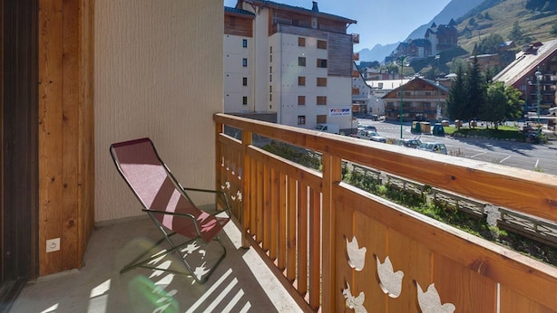 A balcony with a wooden railing, a chair, and a view of a town with buildings and mountains in the background. - Snowfront Stay for Four Guests (Les Deux Alpes)