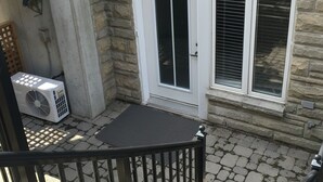 Um edifício de pedra com porta e janelas brancas, uma pequena área externa com um tapete e um aparelho de ar condicionado.