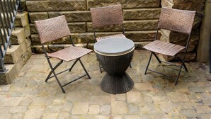 Conjunto de mesa e cadeiras de vime em frente a uma parede de pedra, com uma cerca de metal ao fundo.