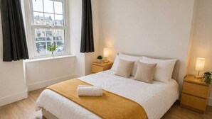 A neatly made bed with a white and brown blanket, two bedside lamps, and a window with curtains. - Central Bristol Apartment with Parking | Sleeps 6 (Bristol City)