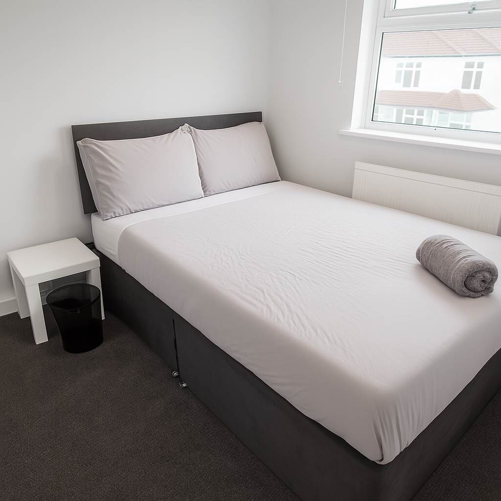 A neatly made bed with a grey pillow, a white bedside table with a black trash bin, and a window with a view of a building.