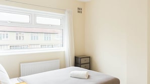 Quarto com cama, mesa de cabeceira, abajur e janela com vista para prédios.