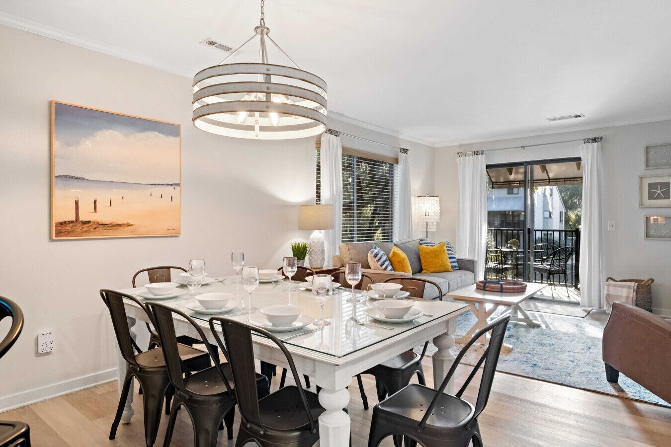 A dining area with a glass table, metal chairs, a chandelier, a painting, and a view to the outside.