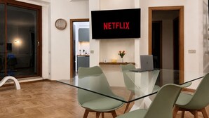 Un salon moderne avec une table en verre, des chaises vertes, un téléviseur à écran plat et une cuisine en arrière-plan.