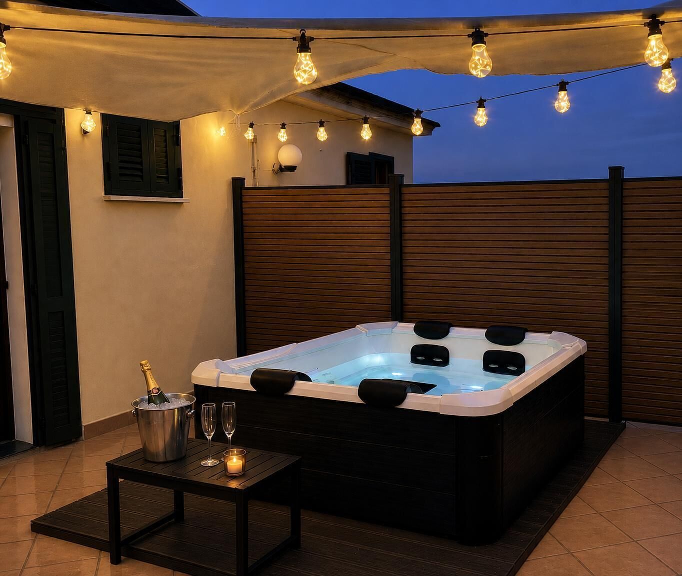 A hot tub with a wooden deck, a table with a bucket of champagne, and string lights.
