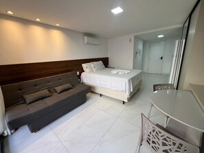 A modern hotel room with a sofa bed, a separate bed, a dining table, and an air conditioning unit. - Unity 223 · Beachfront apartment in Cabo Branco for 3 people (João Pessoa)