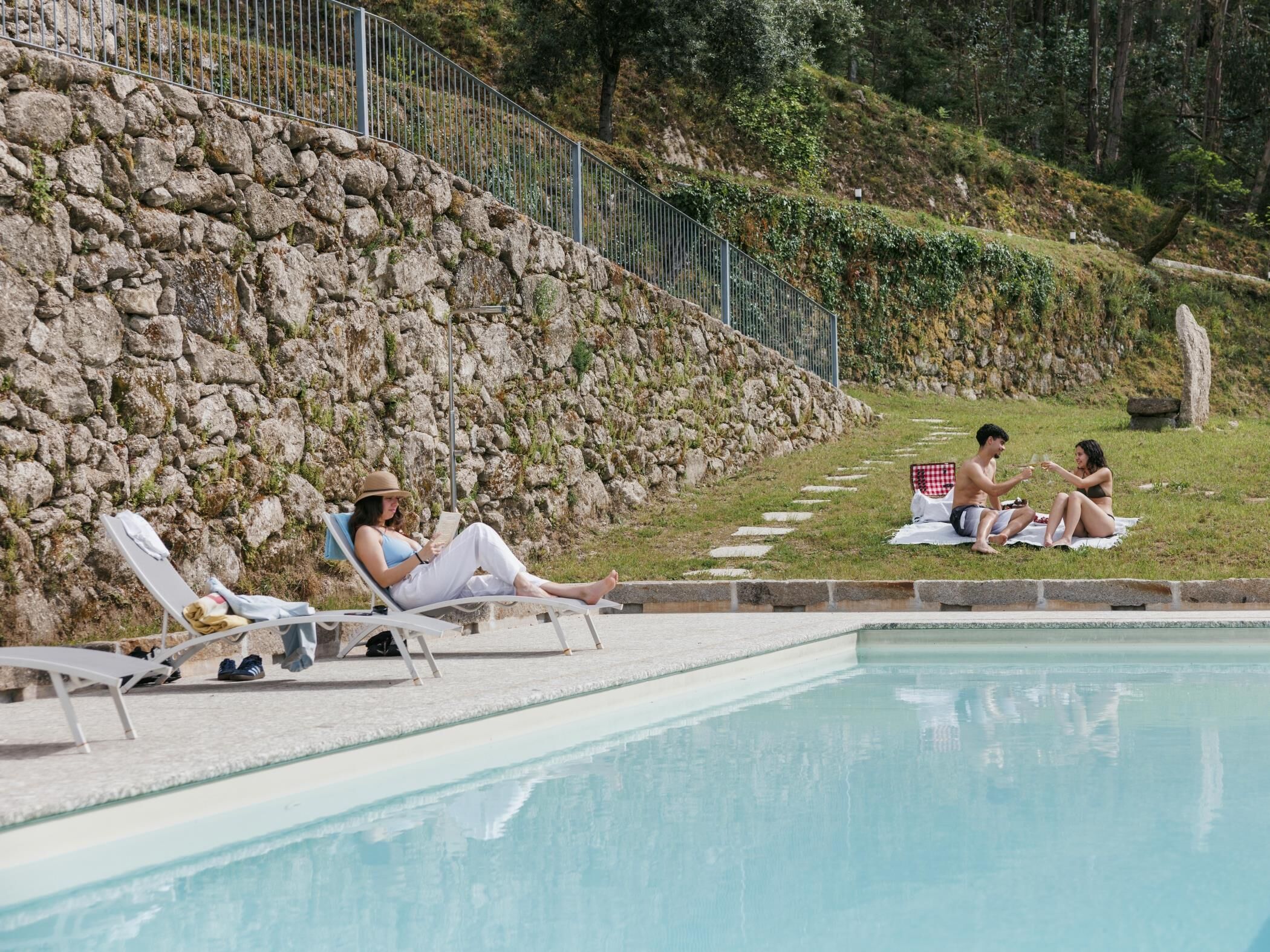 Zona junto a la piscina con sillones de descanso, un muro de piedra y una pendiente cubierta de césped.