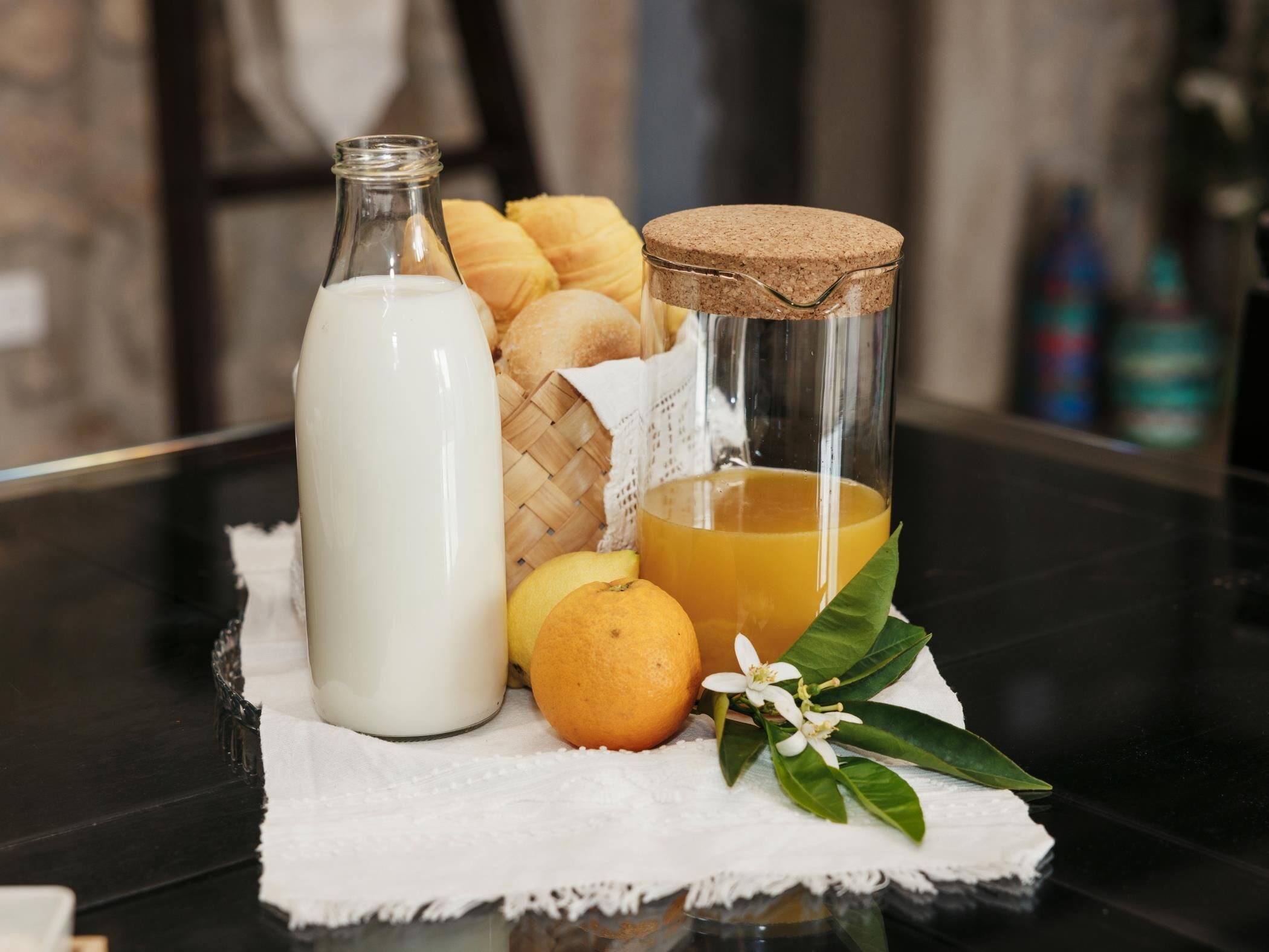 A glass jar with a cork stopper, a bottle of milk, a loaf of bread, an orange, and a sprig of greenery on a table.