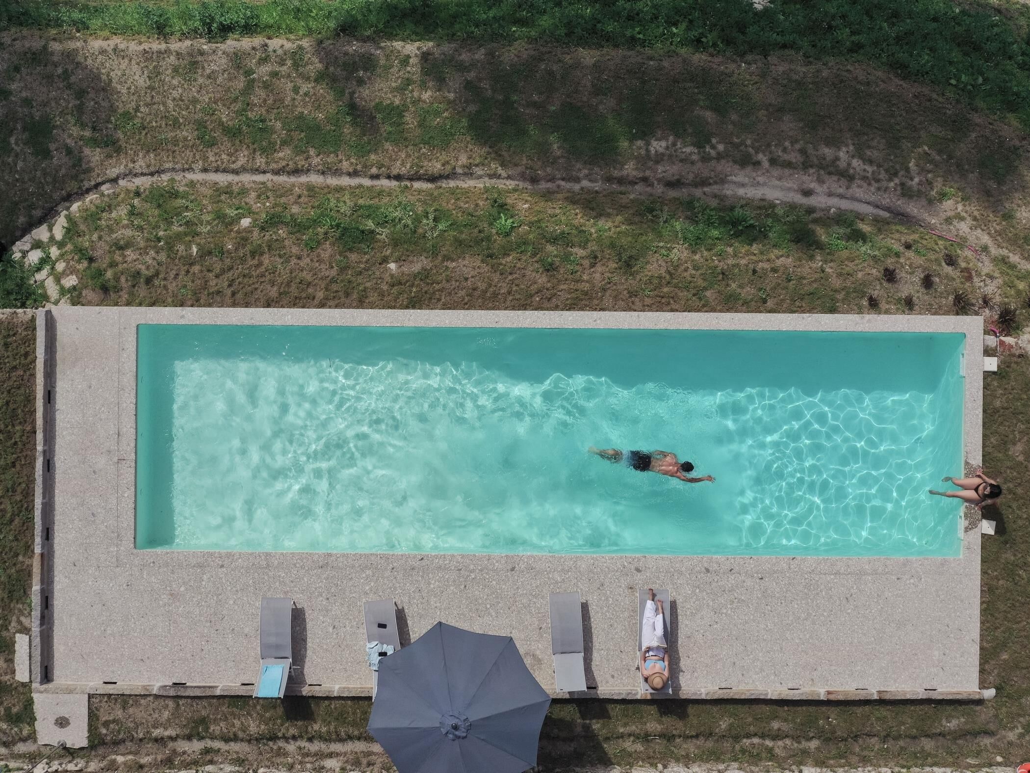 An aerial view of a rectangular swimming pool with clear water, surrounded by a concrete deck with lounge chairs and an umbrella.
