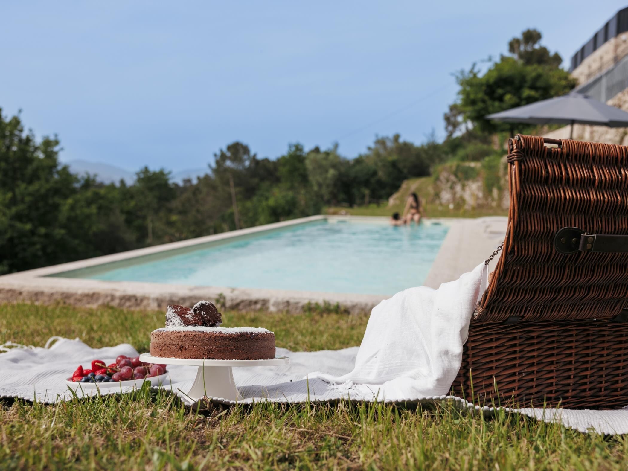Un décor en bord de piscine avec une chaise en osier, un gâteau sur un présentoir et une couverture sur l’herbe.