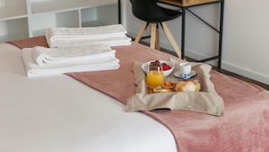 En snyggt bäddad säng med prydligt vikta vita handdukar, ett frukostbricka med croissanter, frukt och apelsinjuice, och ett skrivbord i trä med en svart stol i bakgrunden.
