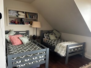 Deux lits jumeaux avec une literie à carreaux, une bibliothèque remplie de livres et d’objets décoratifs, et une lampe posée sur le sol.