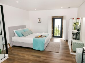 Ein modernes Schlafzimmer mit Bett, einem kleinen Tisch mit Frühstücksangebot, einem Badezimmer mit Waschbecken und Spiegel sowie einem Balkon mit Pflanzen.