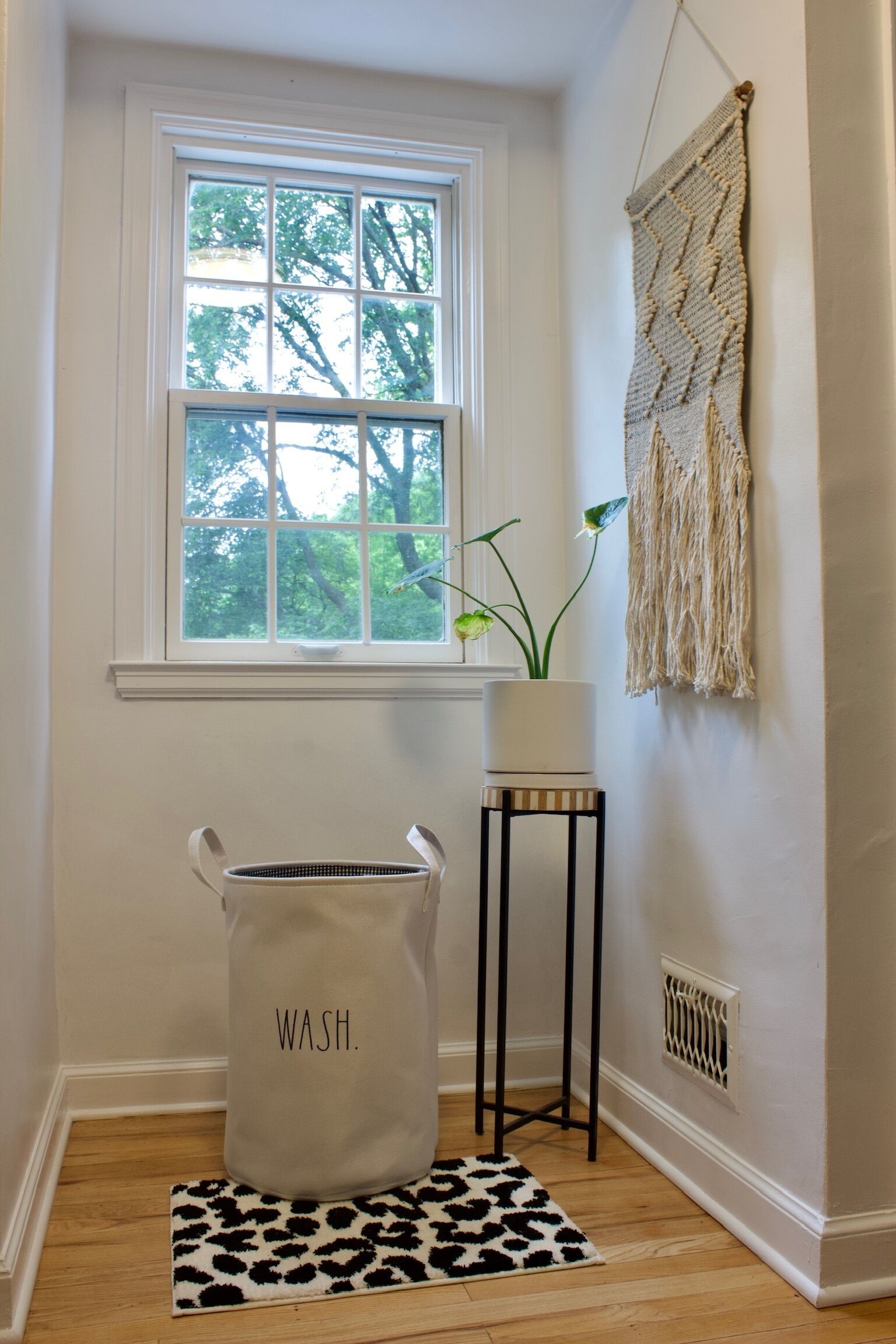 A laundry basket labeled 'WASH.' in a room with a window, a plant, and a macramé wall hanging.