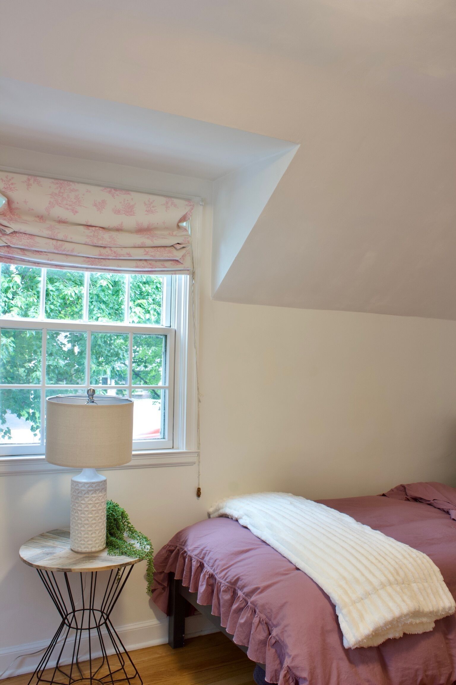 Un dormitorio con ventana, lámpara, cama con ropa de cama rosa y un plaid blanco.