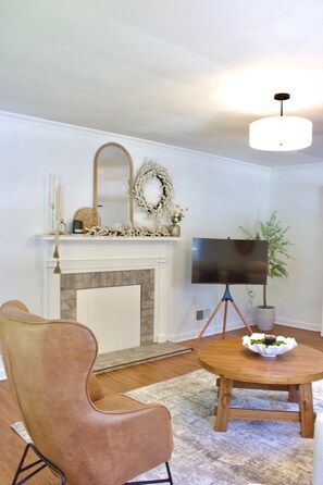 A living room with a fireplace, a flat-screen TV on a stand, a round wooden table with a bowl, and a brown leather chair.
