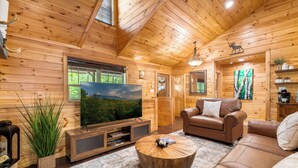 Ein gemütliches Blockhaus-Interieur mit Fernseher, Ledersofa und großem Fenster mit Blick auf Grünflächen.