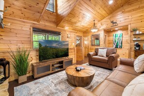 Ein gemütliches Blockhaus-Interieur mit Fernseher, Ledersofa und großem Fenster mit Blick auf Grünflächen.