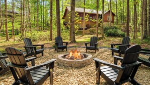 Ein kreisförmiger Feuerkorb, umgeben von Adirondack-Stühlen, in einer Waldlandschaft mit einer Holzhütte im Hintergrund.