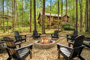 Ein kreisförmiger Feuerkorb, umgeben von Adirondack-Stühlen, in einer Waldlandschaft mit einer Holzhütte im Hintergrund.