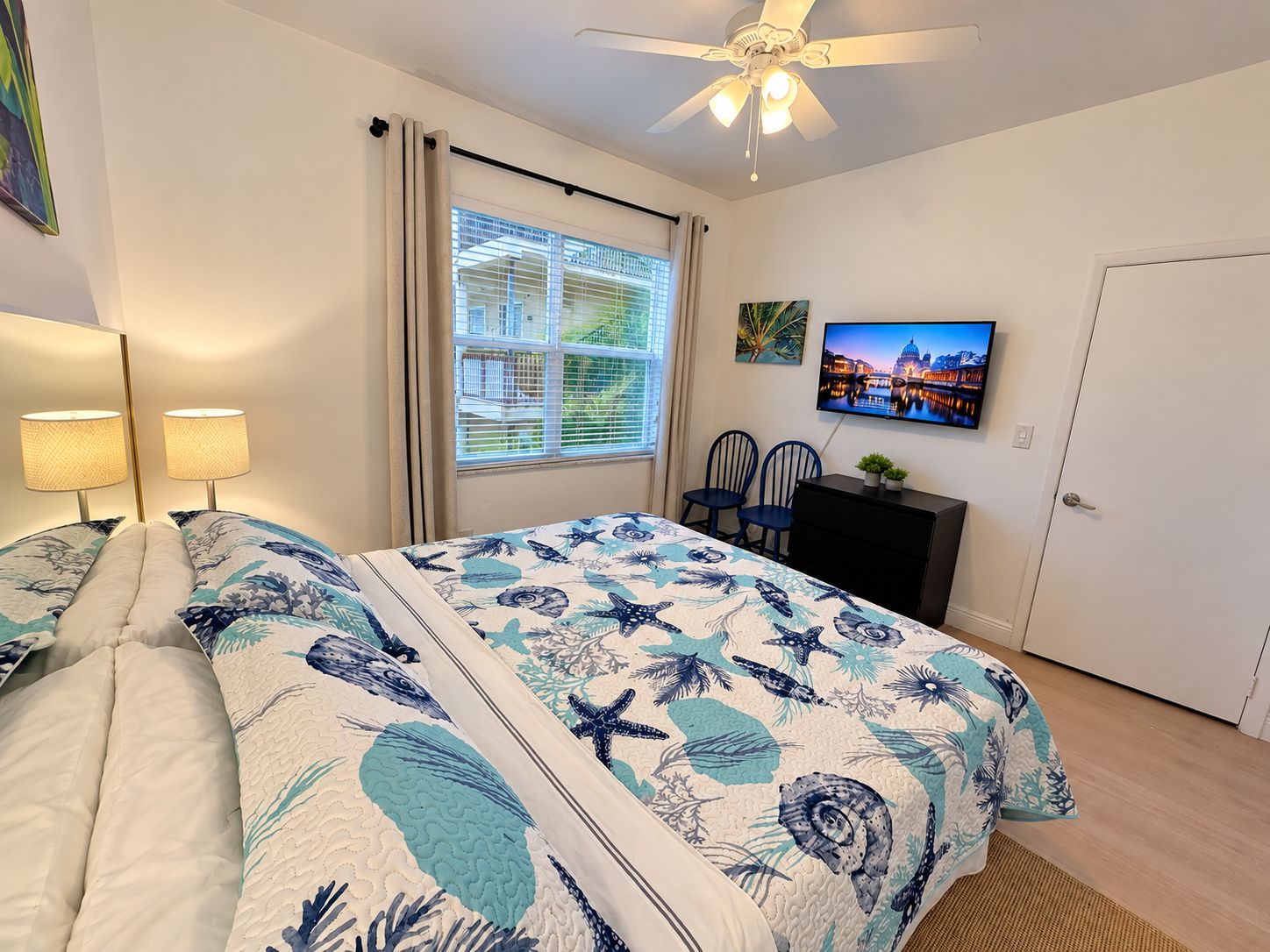 Un dormitorio con cama, ventilador de techo, ventana con persianas, televisor en la pared y una puerta.