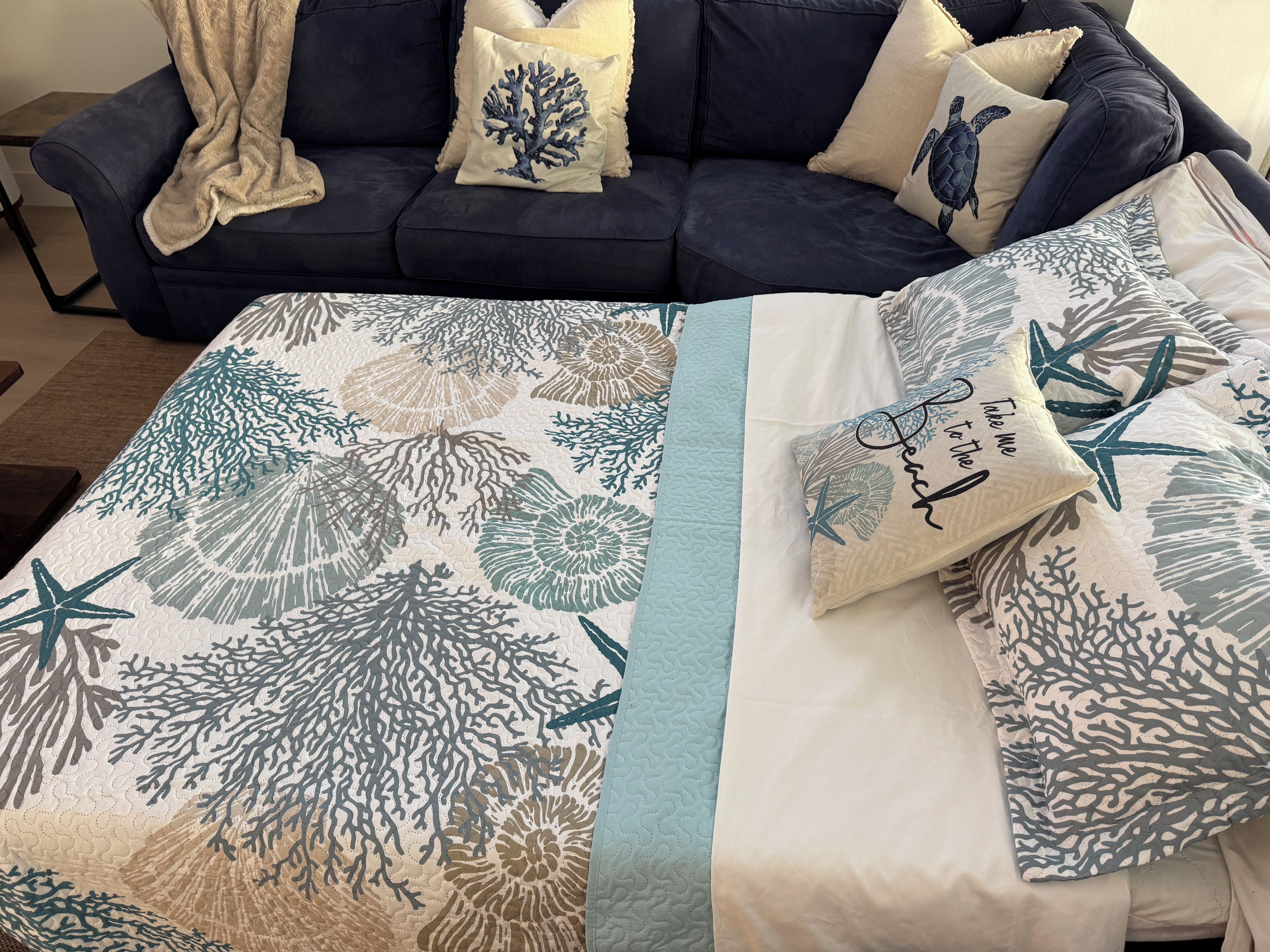 A bed with a coral print bedspread and pillows, a dark blue sofa with decorative pillows, and a blanket draped over the sofa.