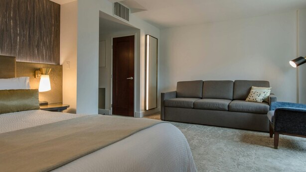 Una habitación de hotel con una cama, un sofá, un escritorio con una lámpara y una puerta que da a otra habitación.