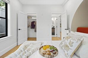 Un dormitorio con cama, mesita de noche con lámpara, ventana y armario con ropa y cesta.
