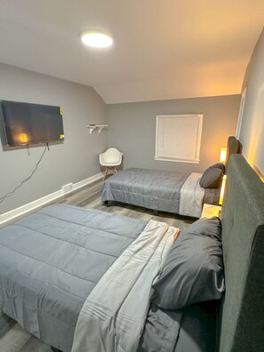 A bedroom with two beds, a TV mounted on the wall, a window with blinds, and a chair. - 3500sf, Arcade, 6bdrm,  2 kitchen, 3baths, Parking (Niagara Falls)