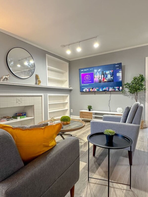 A modern living room with a flat-screen TV mounted on the wall, a round coffee table, a grey sofa with a yellow pillow, and a white shelving unit. - 3500sf, Arcade, 6bdrm,  2 kitchen, 3baths, Parking (Niagara Falls)