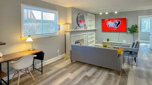 A modern living room with a sofa, a TV, a desk with a chair, and a window with blinds. - Arcade, Close to Fall, 4bdrm,  off street Parking (Niagara Falls)
