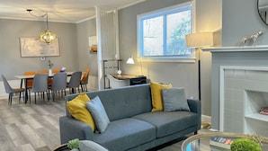 A modern living room with a grey sofa, a dining area with orange chairs, a fireplace, and a large window. - Arcade, Close to Fall, 4bdrm,  off street Parking (Niagara Falls)