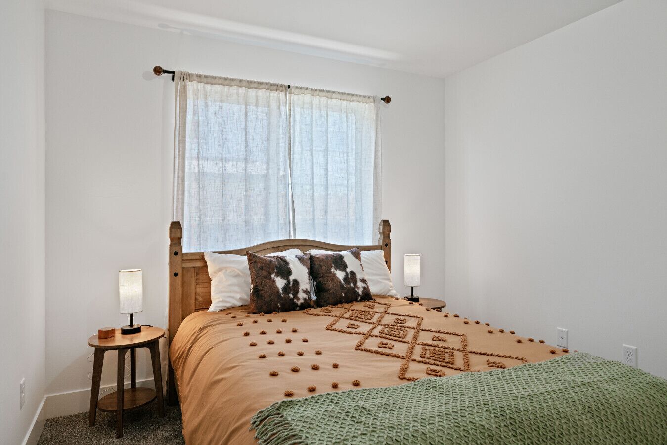 Een slaapkamer met een houten bed, twee bedlampen, een raam met transparante gordijnen en een groene gebreide plaid.