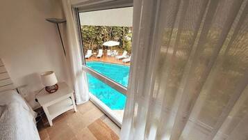 Una habitación con vista a una piscina, una cama, una mesita de noche con lámpara y una ventana con cortinas.