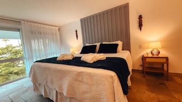 Habitación de hotel con cama, mesita de noche, lámpara y vista a zonas verdes por la ventana.